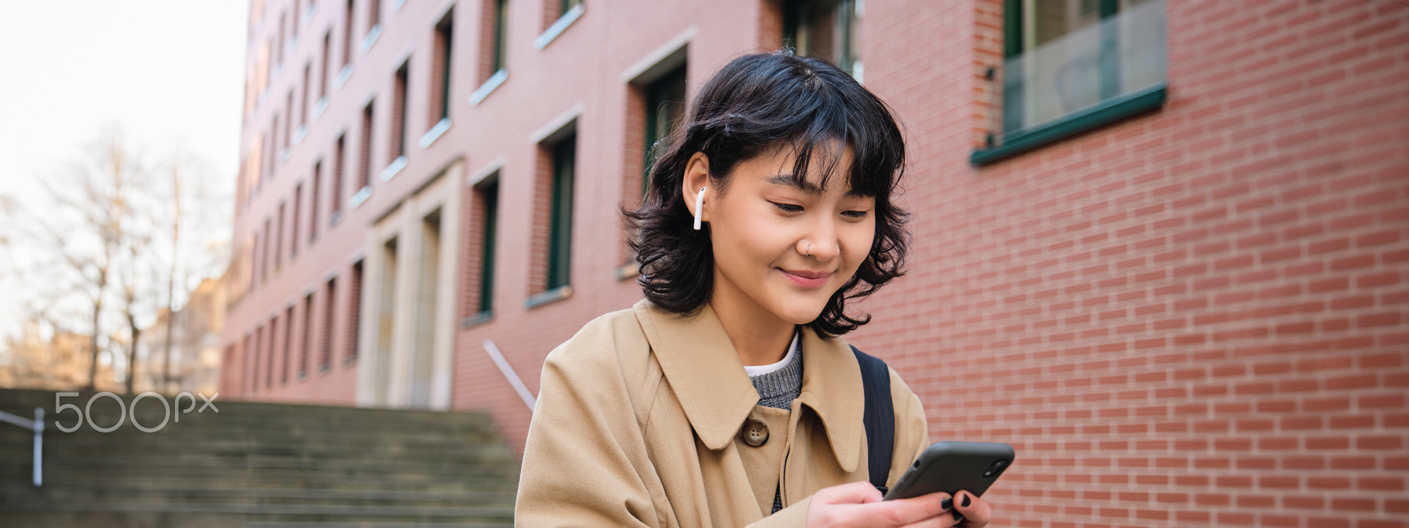 Happy korean girl walks on street, listens music in wireless earphones and holds smartphone, picks