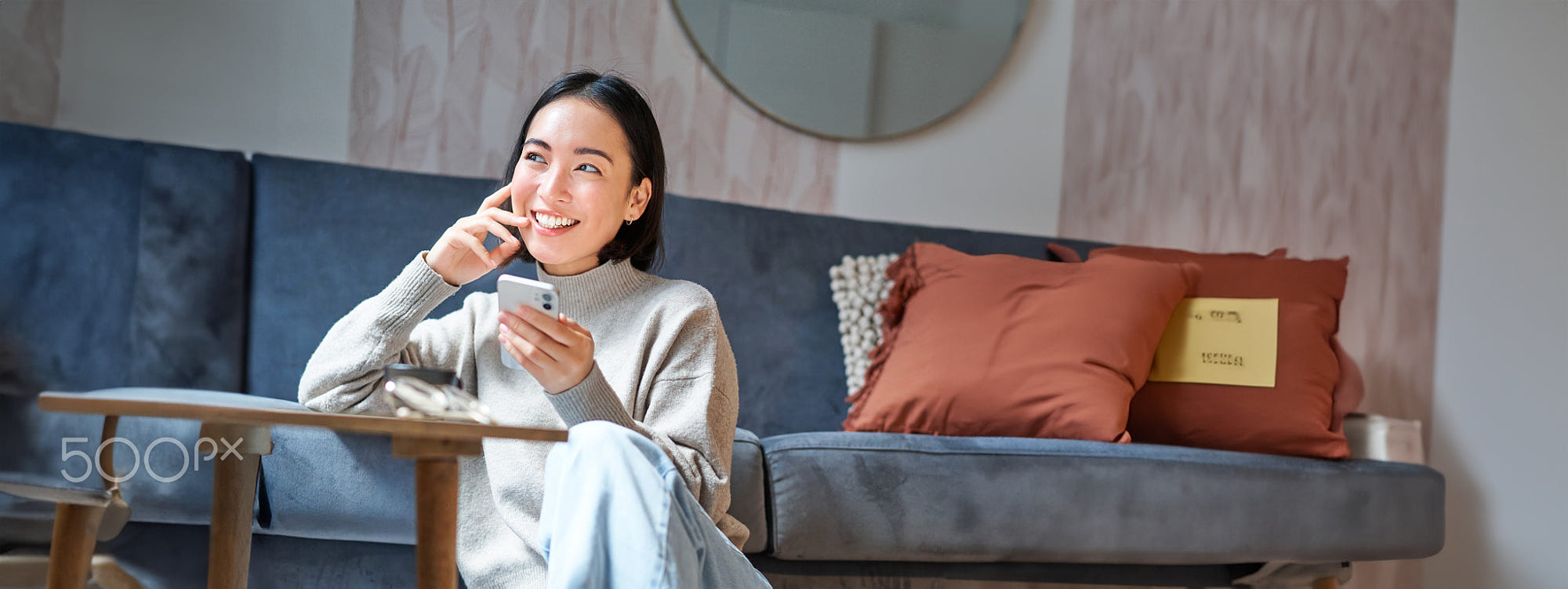 Vertical shot of stylish korean woman sitting on floor at home, using her mobile phone, holding: Vertical shot of stylish korean woman sitting on floor at home, using her mobile phone, holding smartphone and smiling.