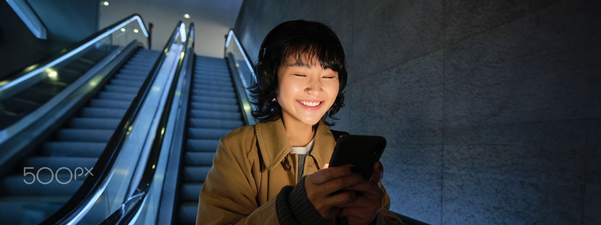 Smiling korean girl going down escalator in dark, holding mobile phone, using smartphone app