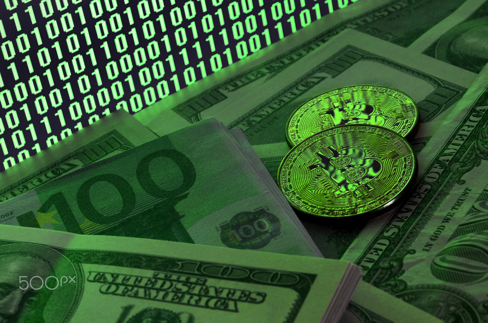 Two bitcoins lies on a pile of dollar bills on the background of a monitor depicting a binary code