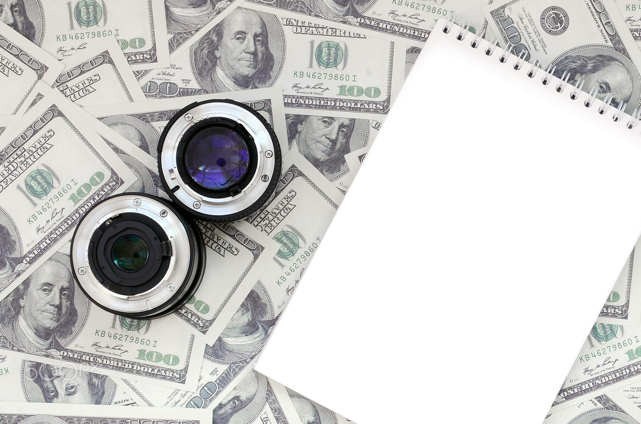 Two photographic lenses and white notebook lie on the background of a lot of dollar bills. Space for