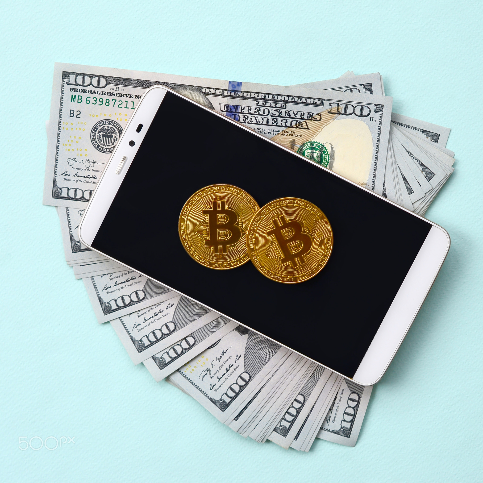 Bitcoins lies on a smartphone and hundred dollar bills on a light blue background