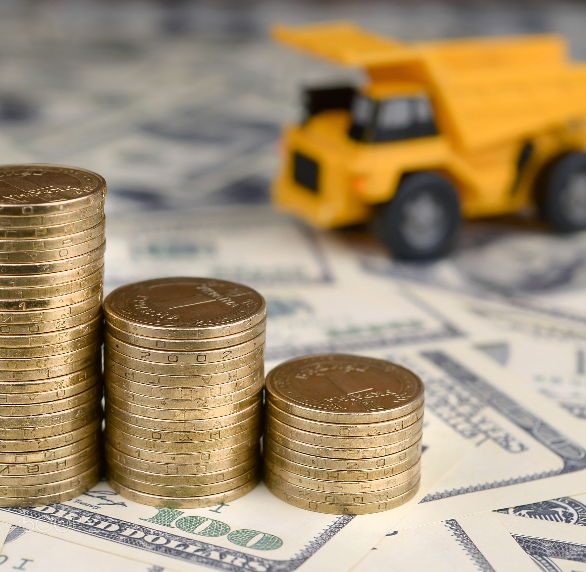 Dump truck toy and stacks of gold coins on background of many hundred dollar bills