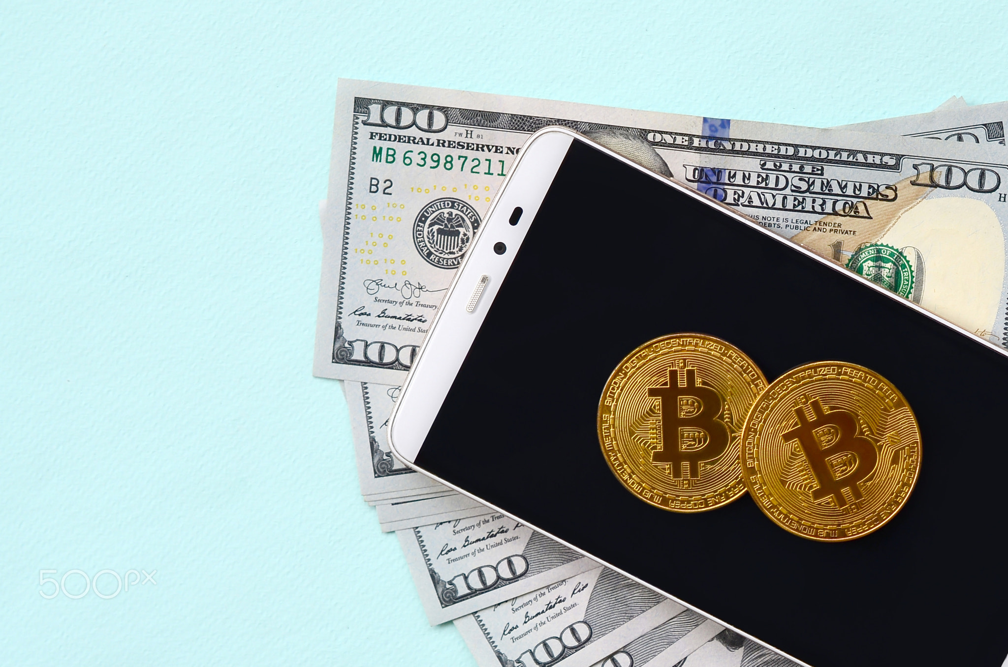 Bitcoins lies on a smartphone and hundred dollar bills on a light blue background