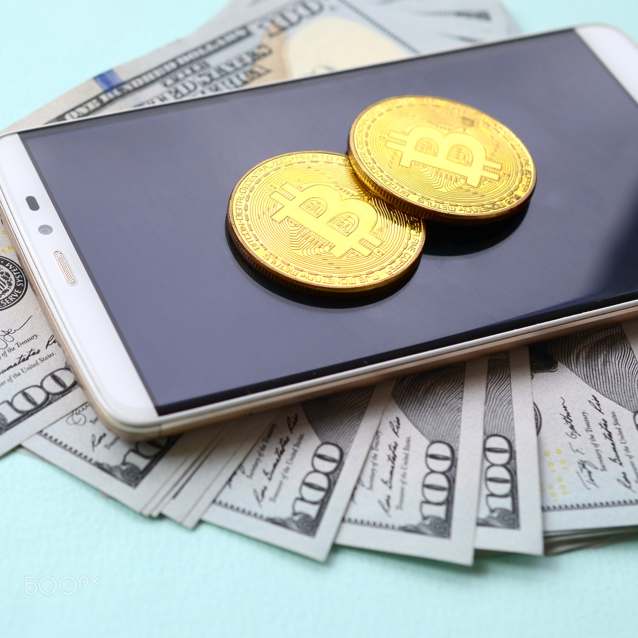 Bitcoins lies on a smartphone and hundred dollar bills on a light blue background