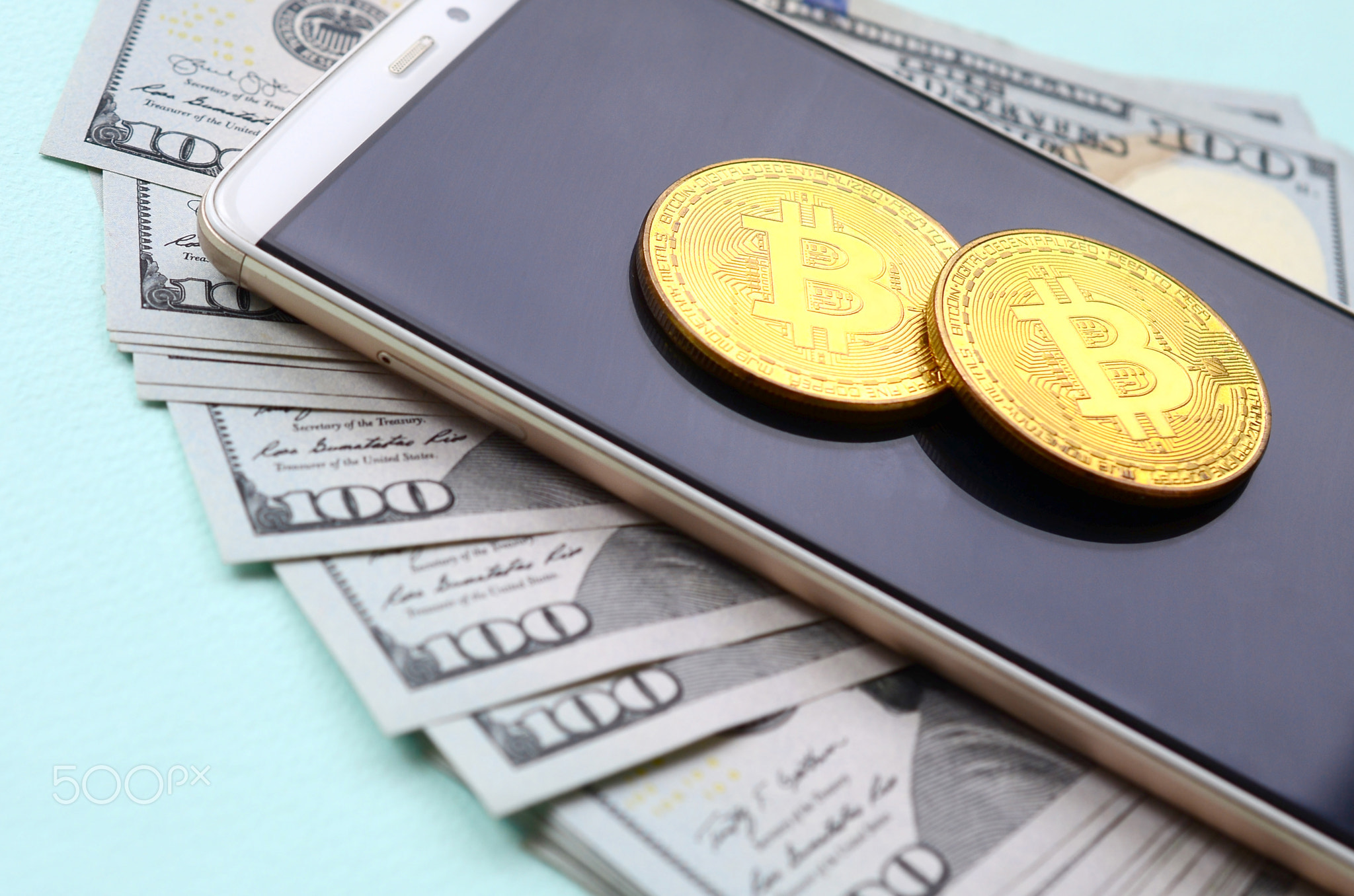 Bitcoins lies on a smartphone and hundred dollar bills on a light blue background