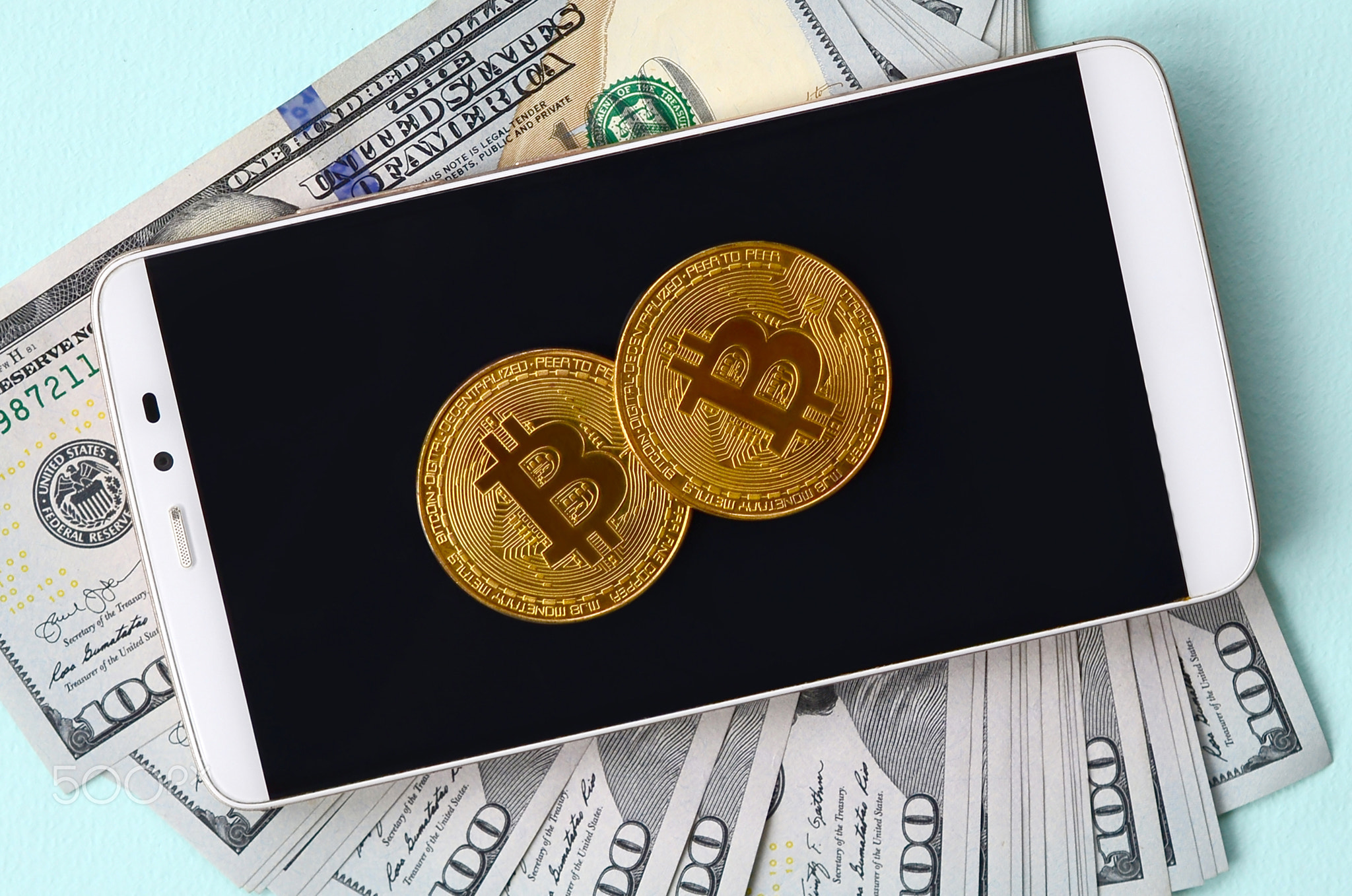 Bitcoins lies on a smartphone and hundred dollar bills on a light blue background