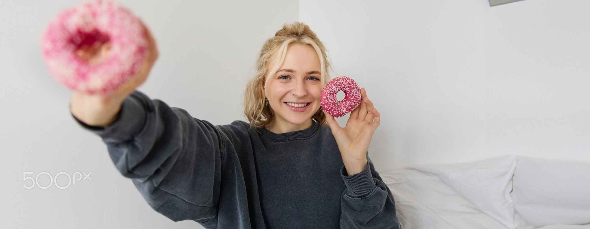 Portrait of beautiful blond woman, showing two pink doughnuts, delicious pastry, eating dessert and