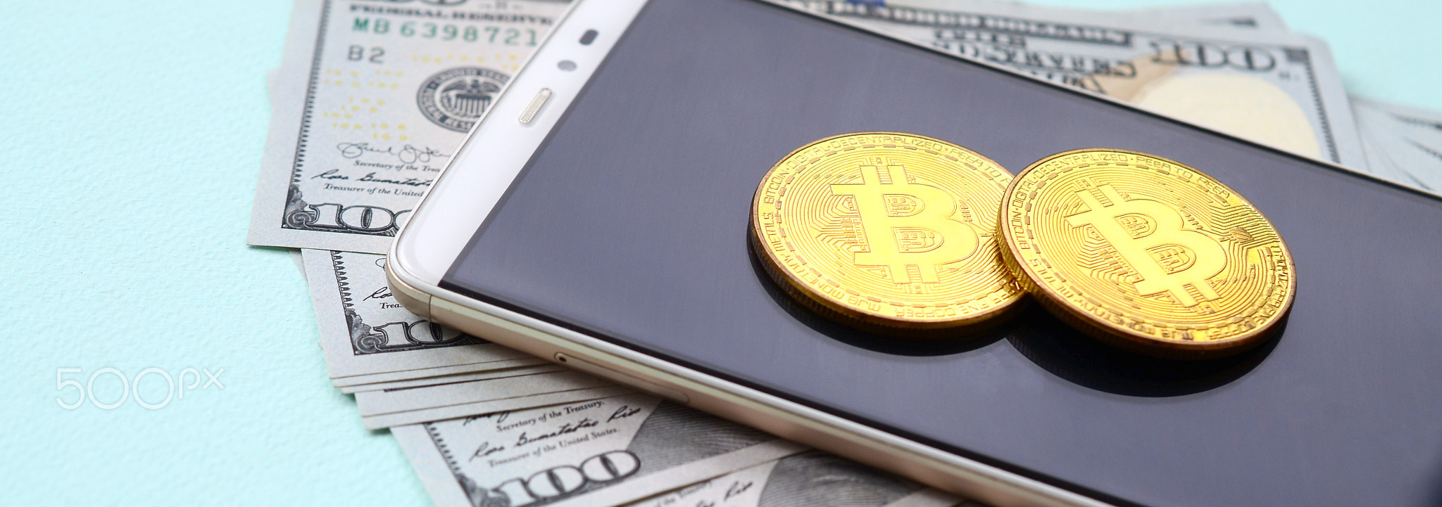 Bitcoins lies on a smartphone and hundred dollar bills on a light blue background