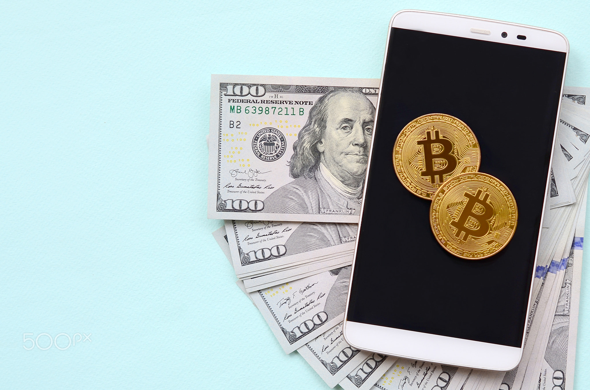 Bitcoins lies on a smartphone and hundred dollar bills on a light blue background