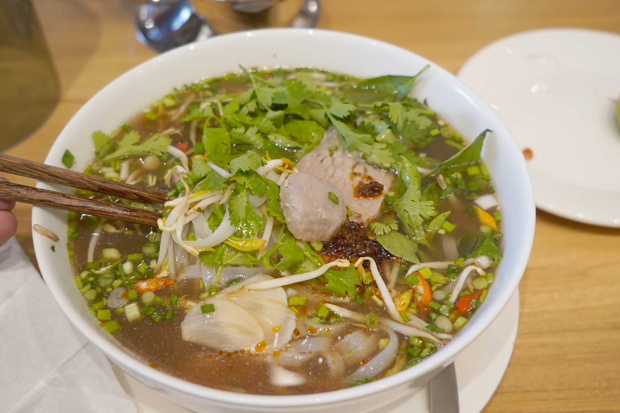 A bowl of pho on a wooden table. The pho appears to have rice noodles, sliced beef, and a clear brot