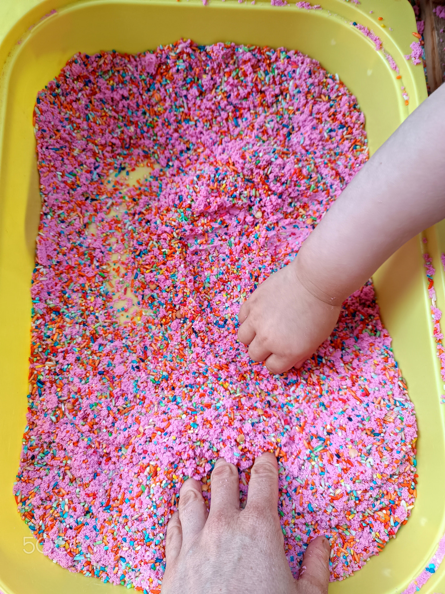 Mammy mother and baby play with touch dyed coloured rice in yellow sensory bin. Sensory play.