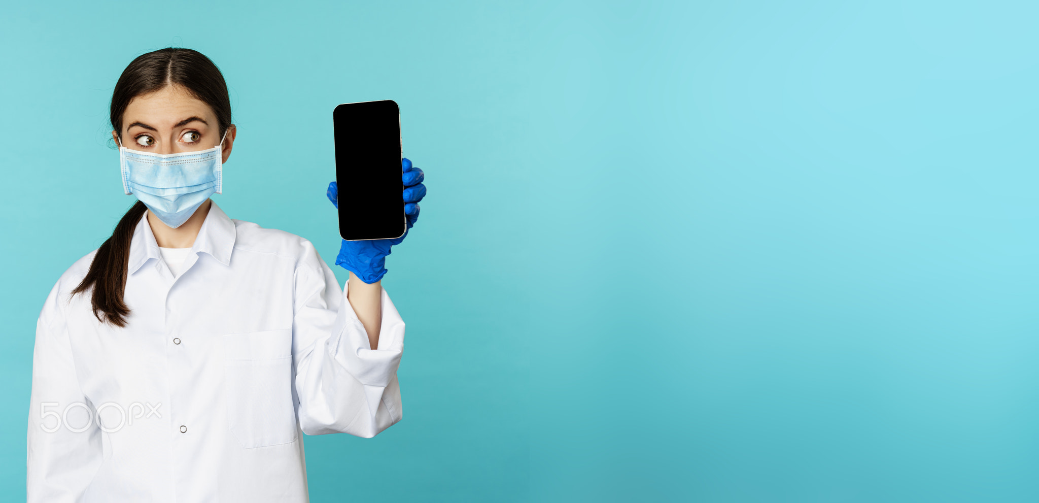 Young woman doctor in medical face mask and hospital uniform, showing mobile phone app, screen