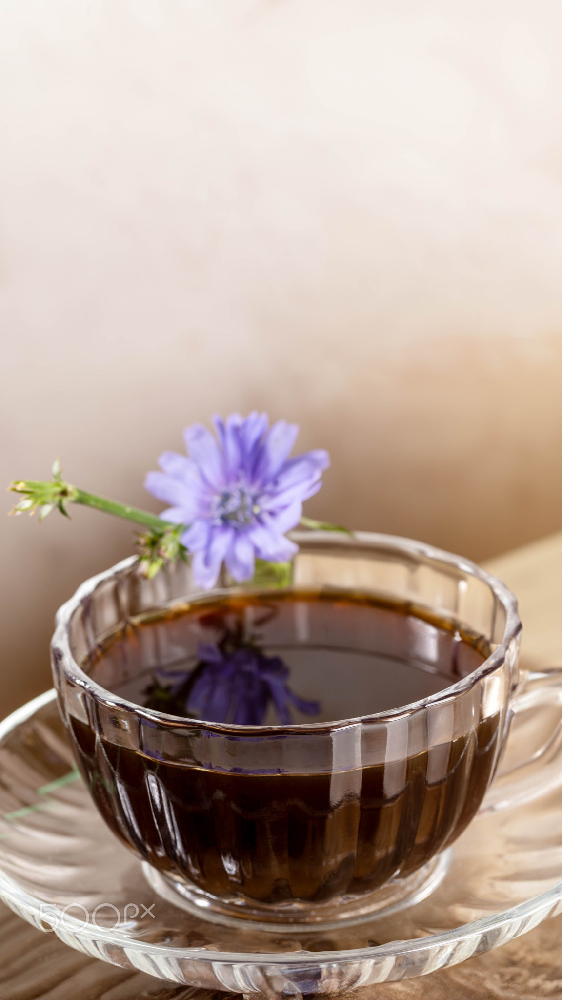 Close up figured glass cup with chicory tea and flower on beige vertical background with copy space.