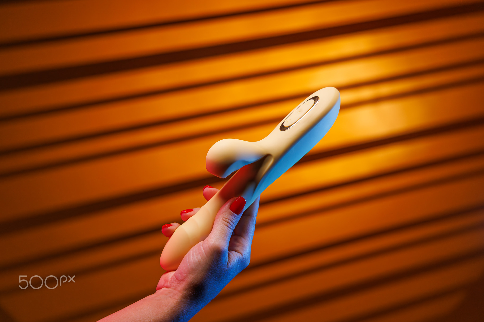 A female hand with red nails firmly holds a yellow vibrator. The background is illuminated by neon l