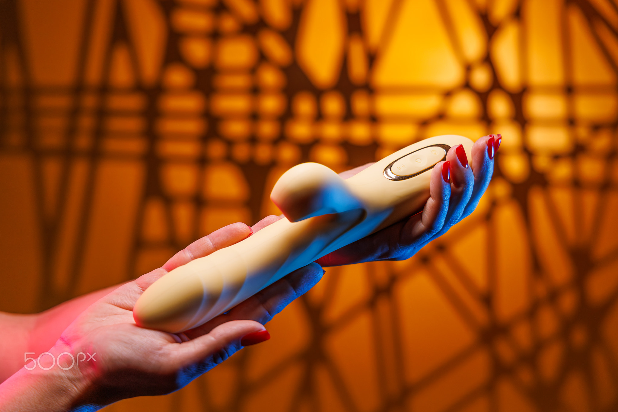 A womans hands with a sexy red manicure rotate a yellow clitoral vibrator. The neon lights and glowi