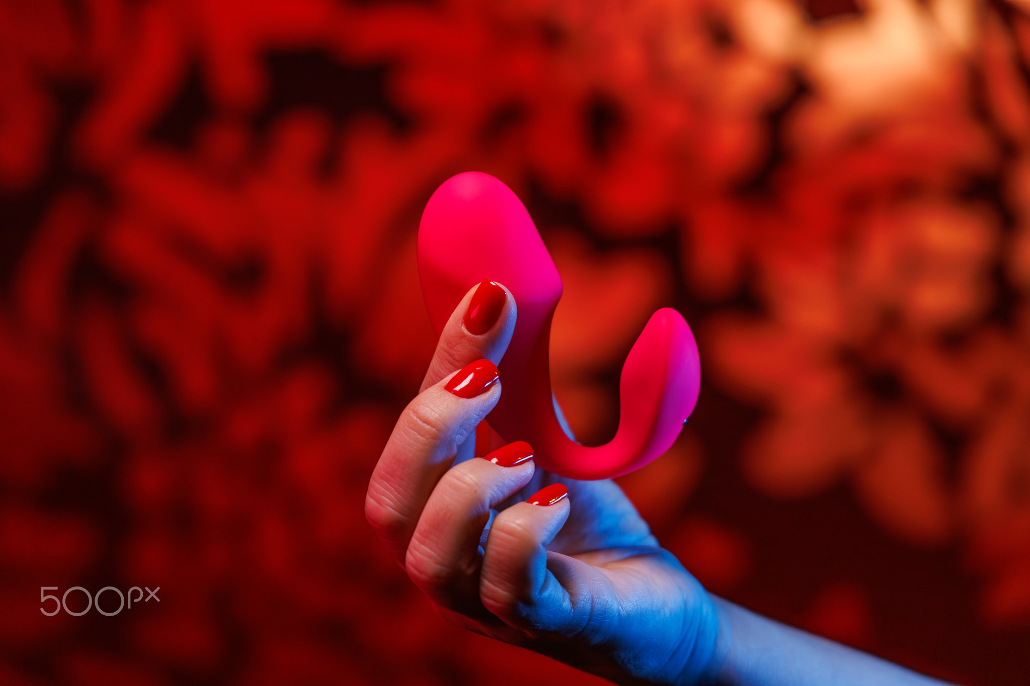 A gentle hand with striking red nails showcases a curved pink vibrator. Neon illumination enhances t