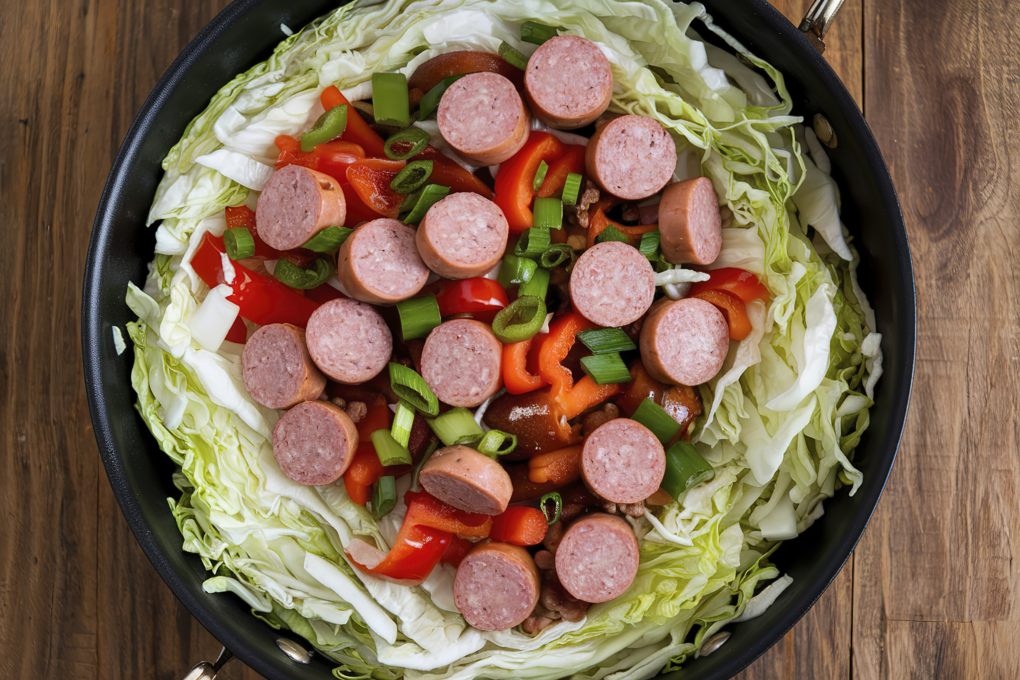 A photo of a skillet filled with shredded cabbage mixed with sliced sausages, diced red and green be