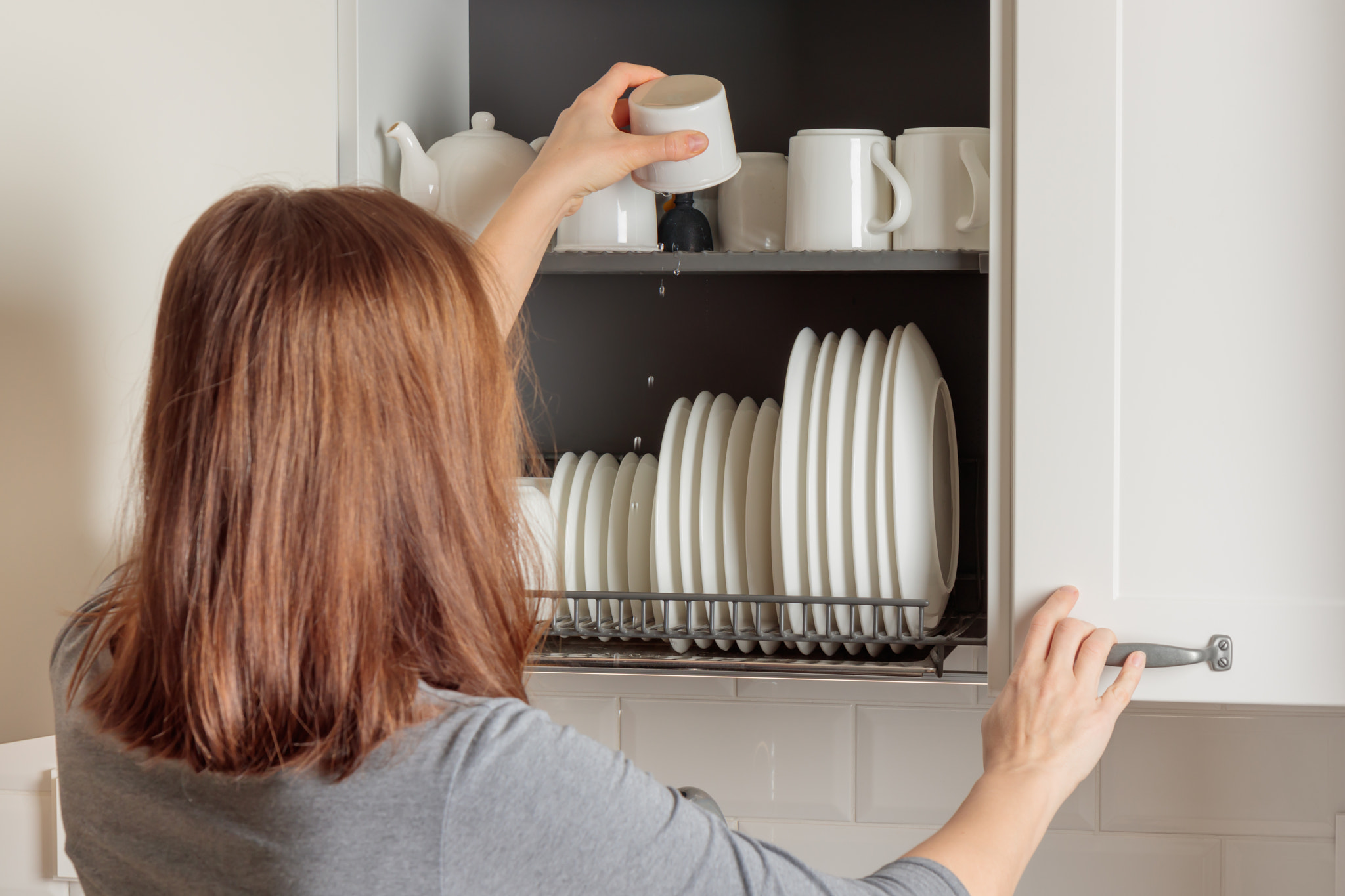 A young woman puts a mug in a dish-drying rack integrated into a wall cabinet in a modern kitchen wi