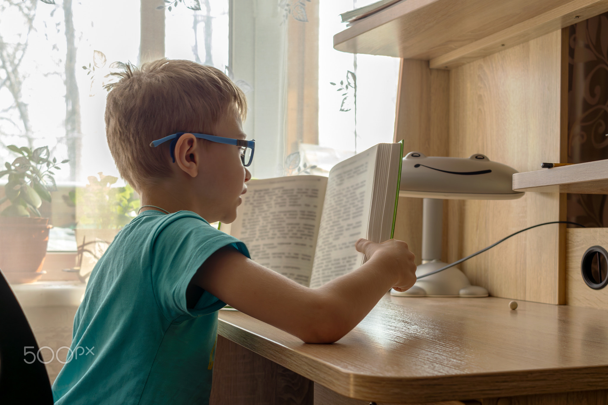 An 8 year old boy wearing glasses is reading book aloud at table by window with indoor plants, lit