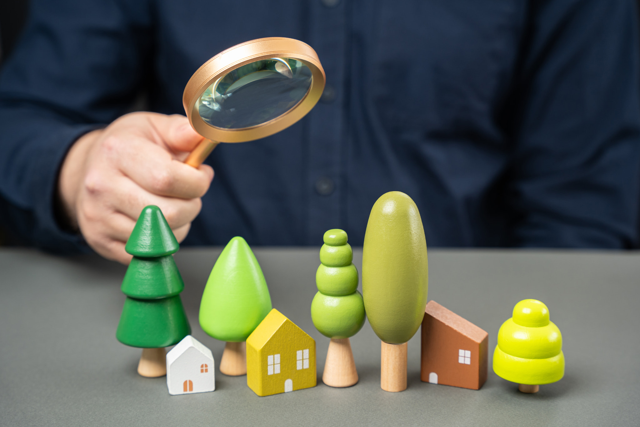 A man holds a magnifying glass over toy trees and houses. Climate and air assessment concept. Scienc