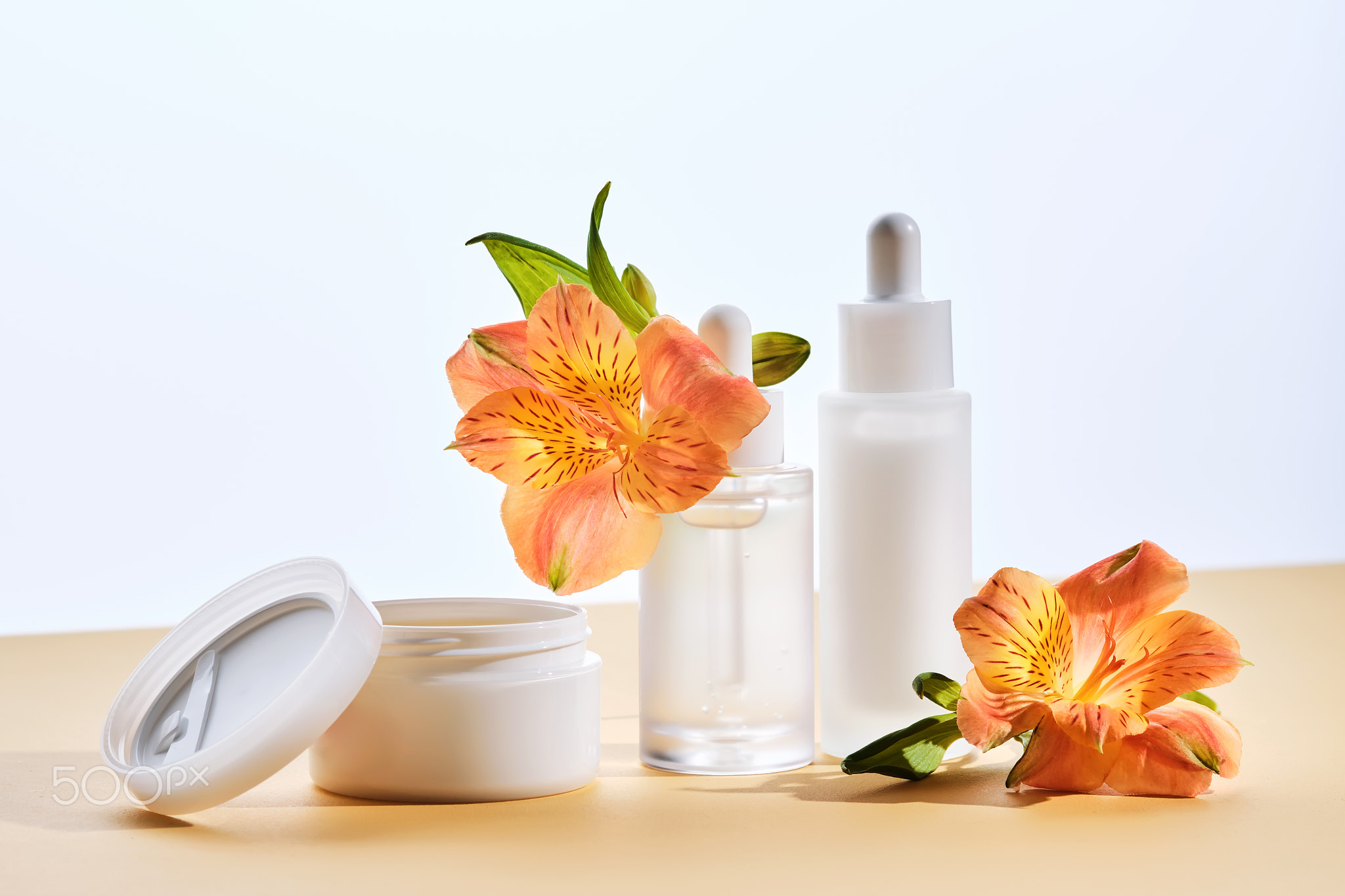Two serums and a cream in a jar with lily flowers on a beige background.