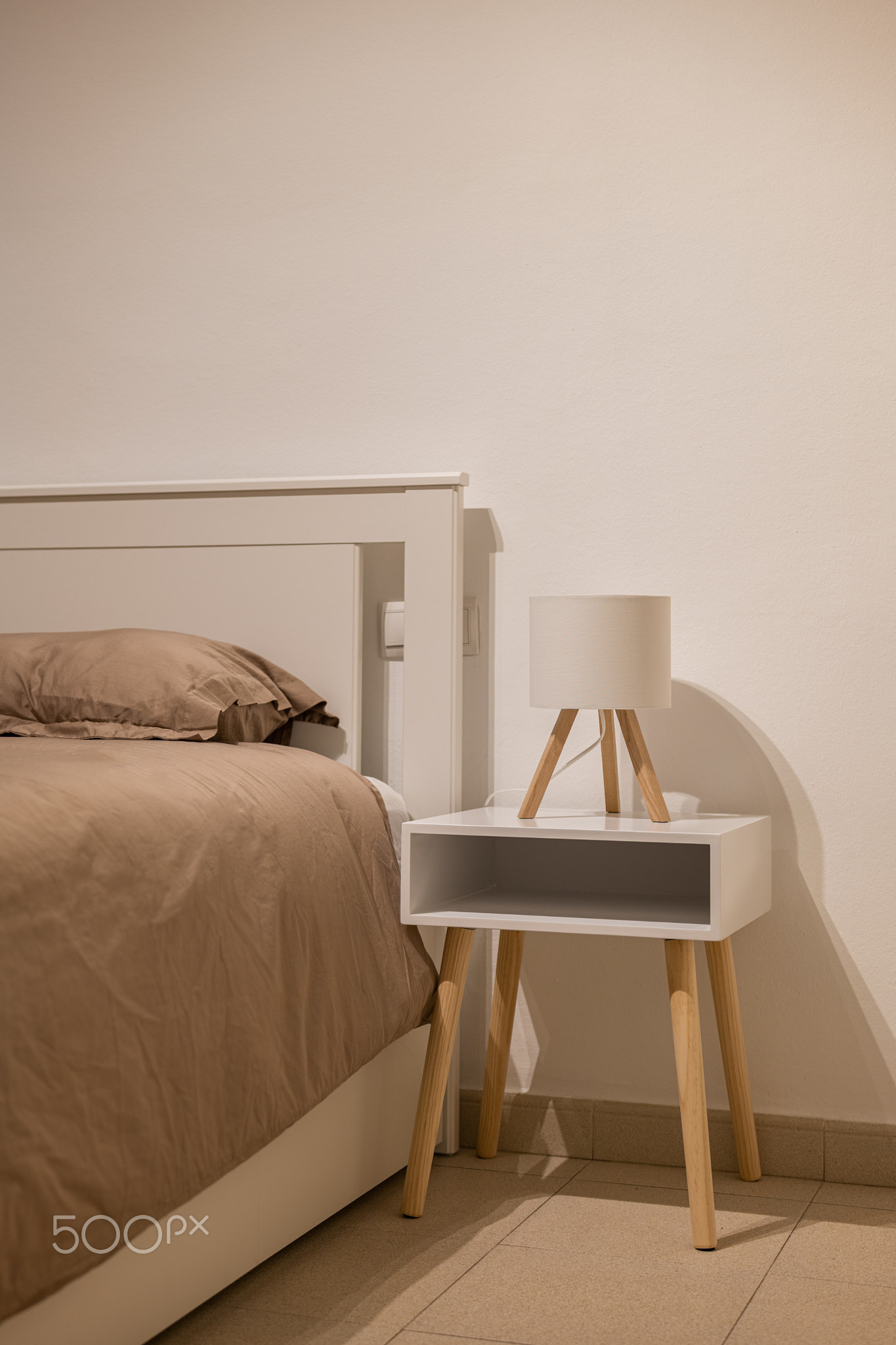 Minimal bedroom corner with taupe bedding, white bed frame and wood nightstand with small lamp