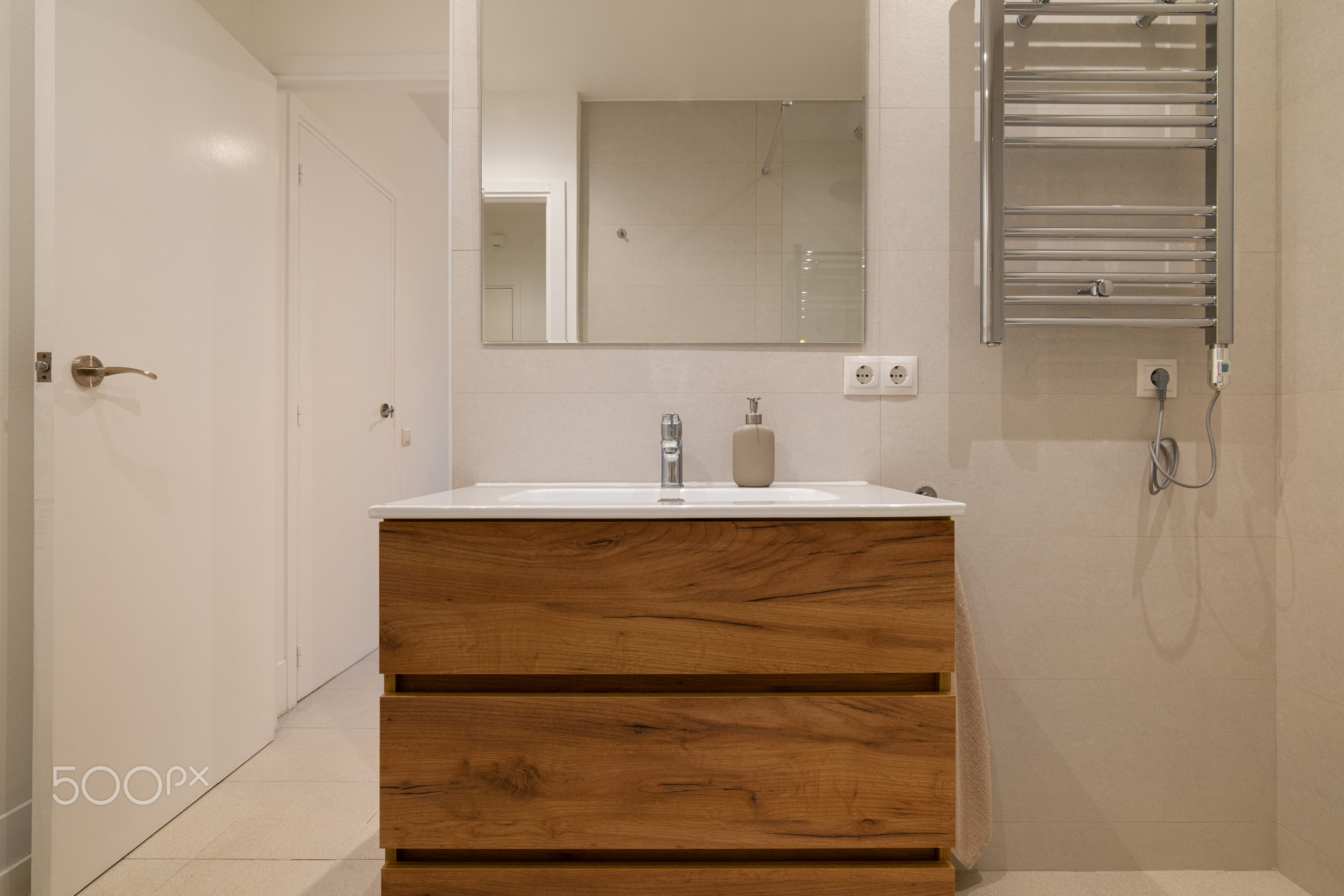 Wide angle of a minimalist bathroom with wood vanity, large mirror, chrome faucet and soap dispenser