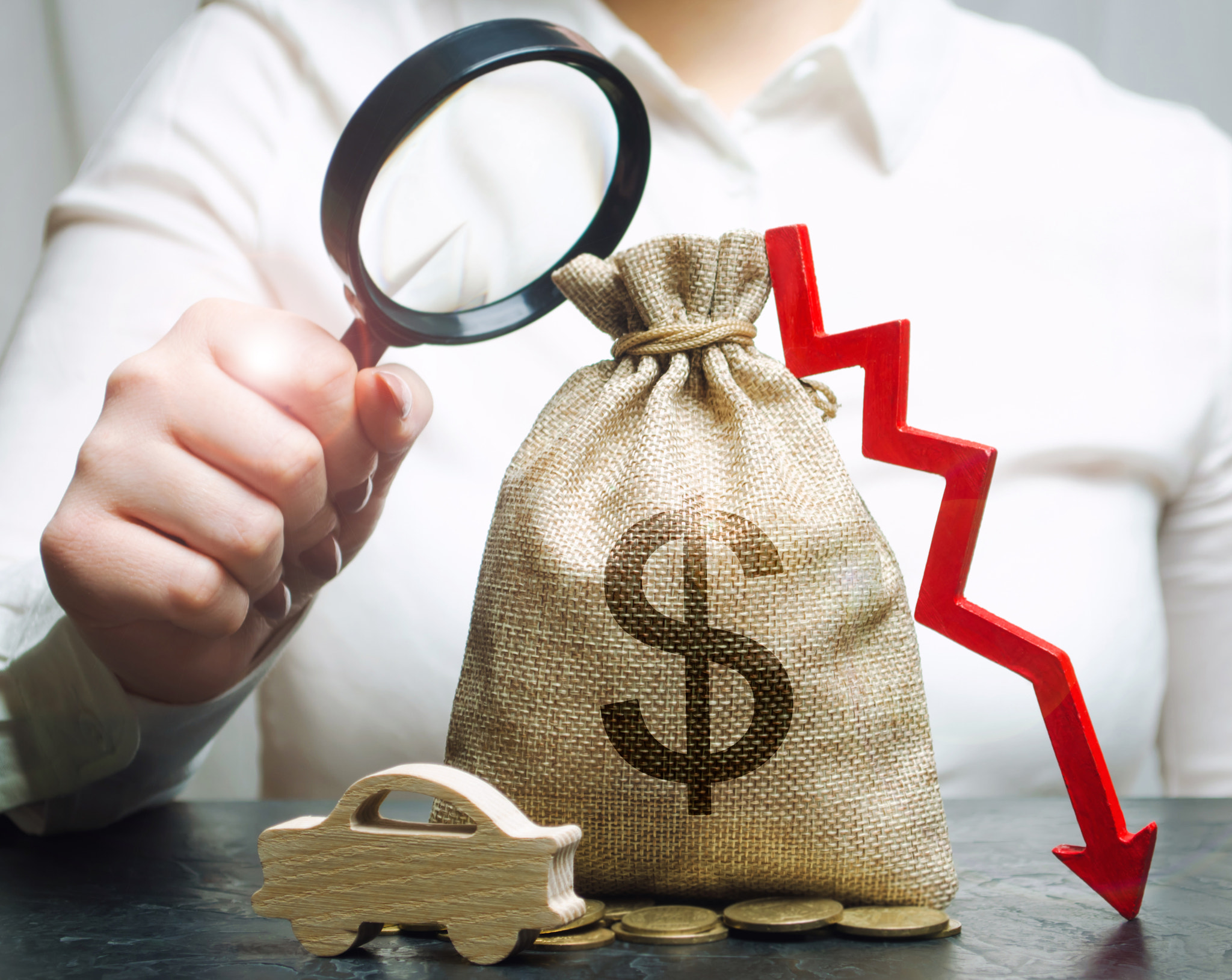 A manager is holding a magnifying glass over a money bag, a car and a down arrow. Falling cost of ma
