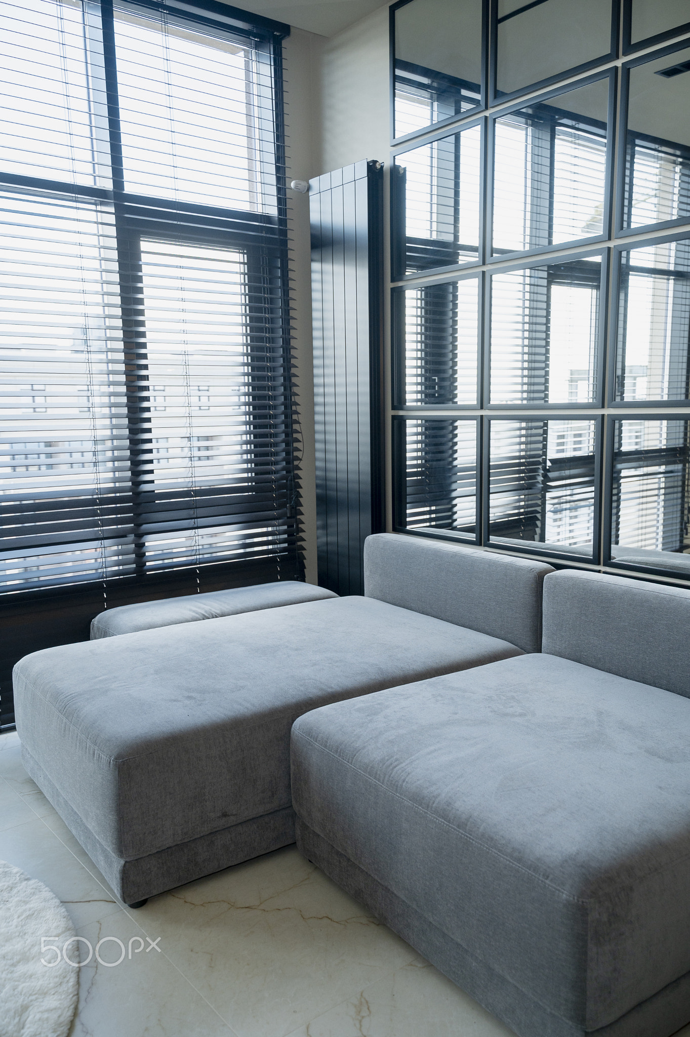 Ergonomic gray sofa standing near a large window with blinds and a mirror with a copy space