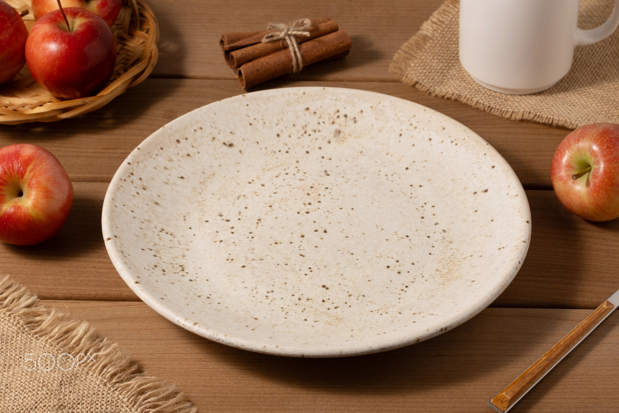 There is an empty plate on a wooden table and red apples nearby