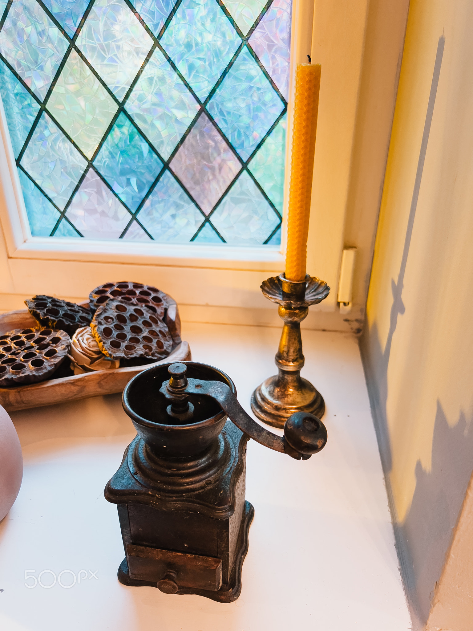 Old coffee grinder resting on windowsill beside beeswax candle and dried lotus pods in wooden bowl