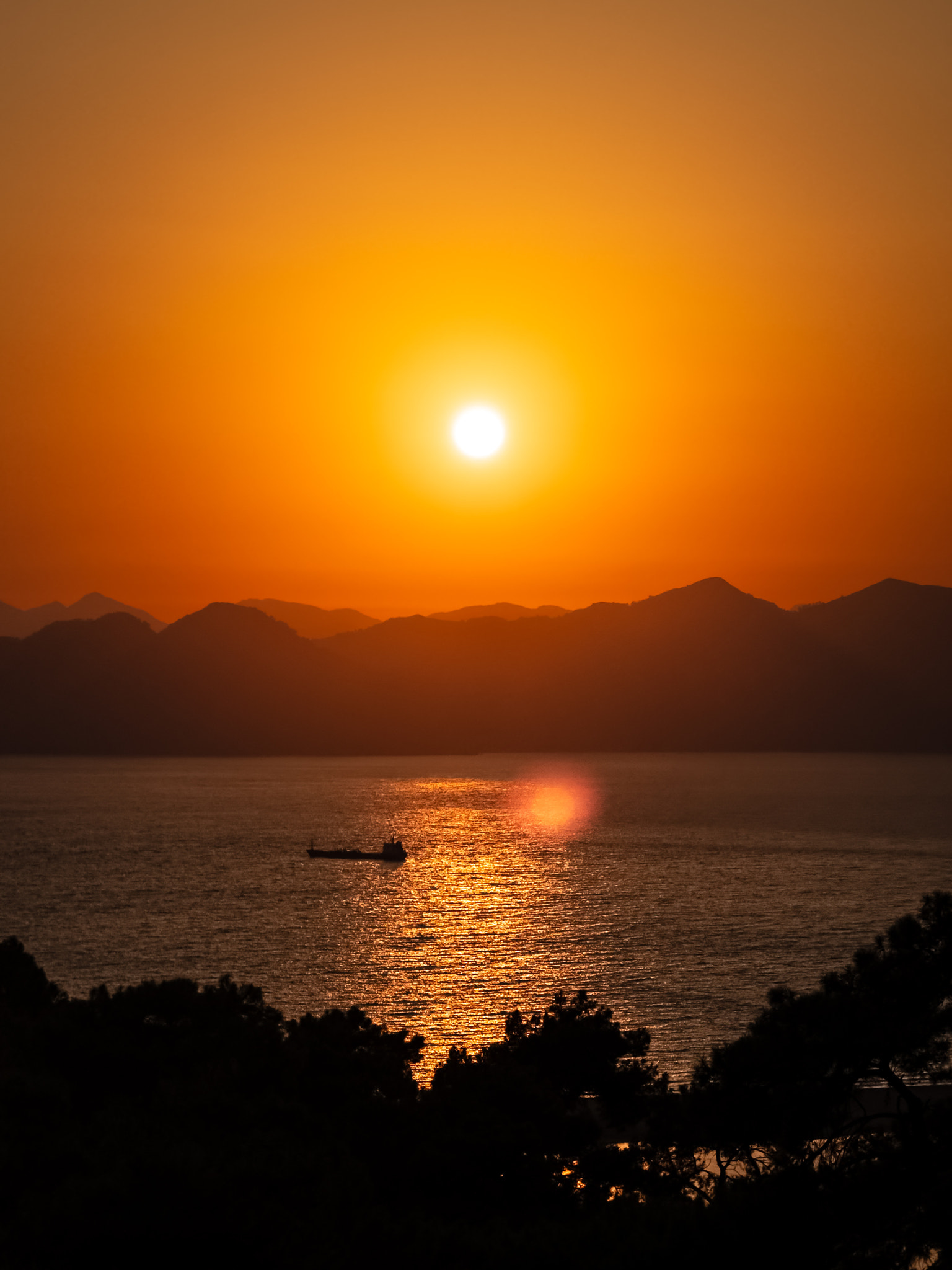 Sun reflecting on Mediterranean sea water at sunset with a cargo ship silhouette and mountain backgr