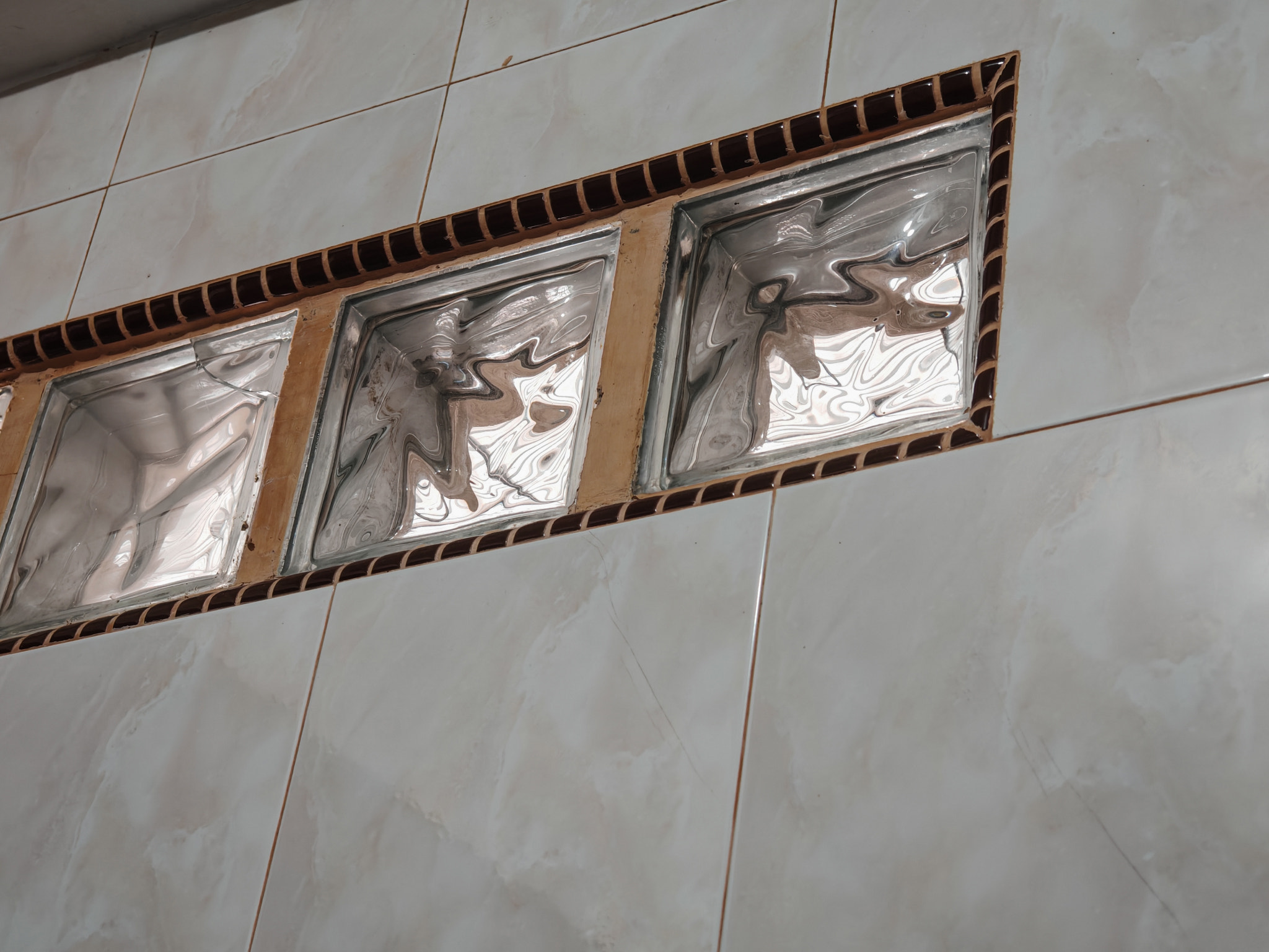 Close-up of a hairline crack in ceramic wall tiles near a glass block window in a modern bathroom