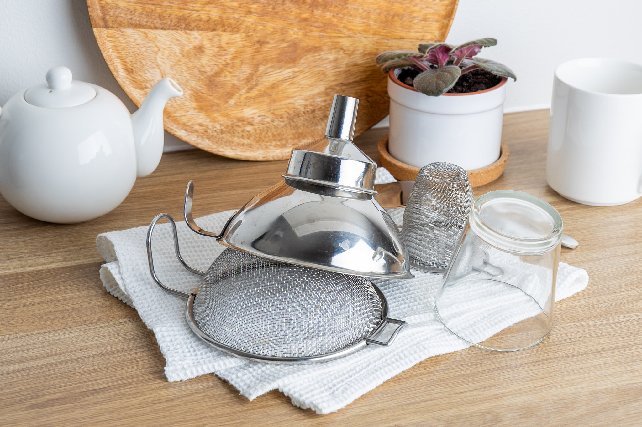 A silver tea strainer sits on a wooden table with a white teapot and a glass jar, ready for use.