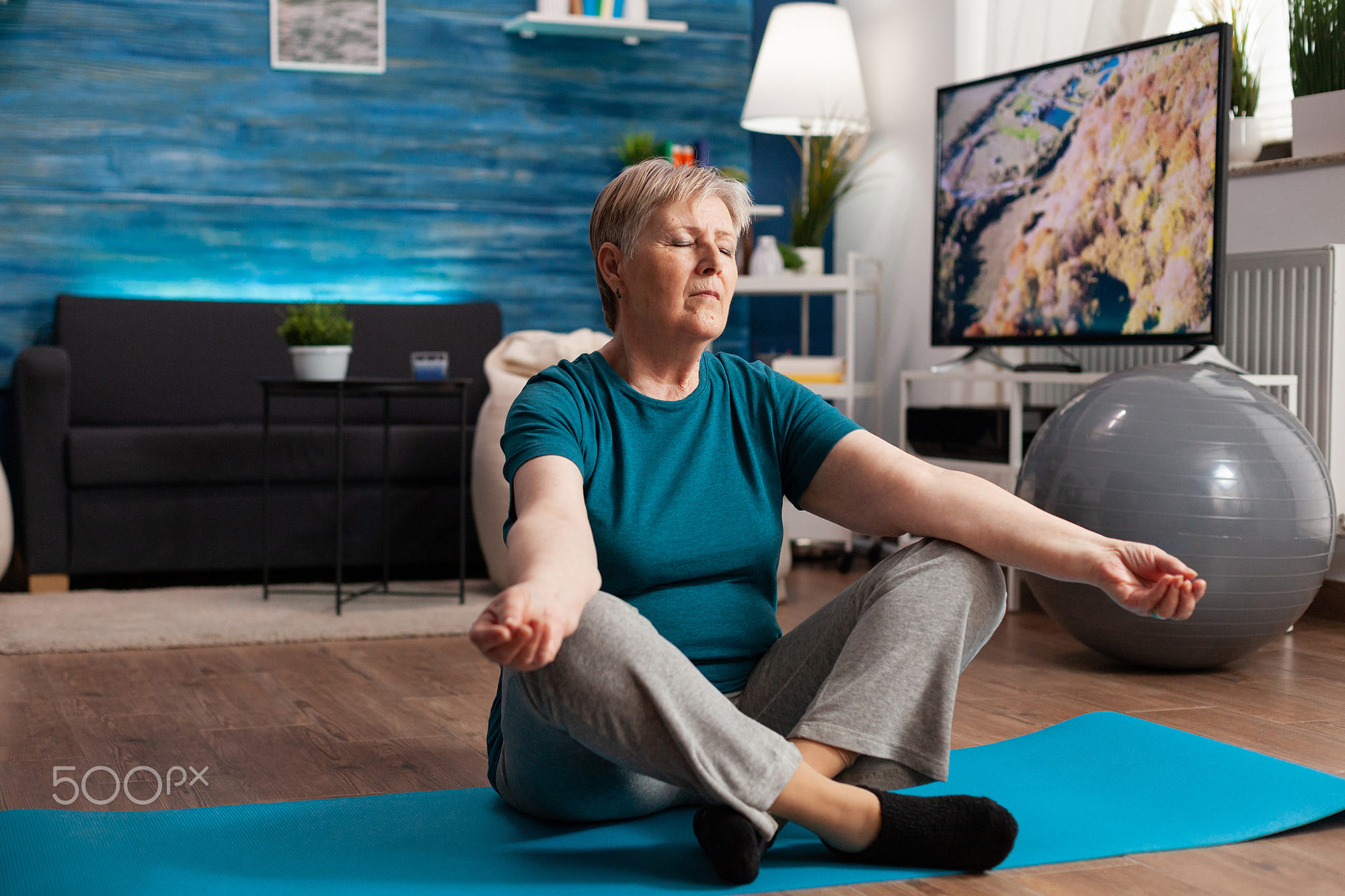Peaceful senior woman with closed eyes sitting on yoga mat meditating during wellness workout: Peaceful senior woman with closed eyes sitting on yoga mat meditating during wellness workout. Comfortable pensioner practicing lotus position exercising body concentration in living room