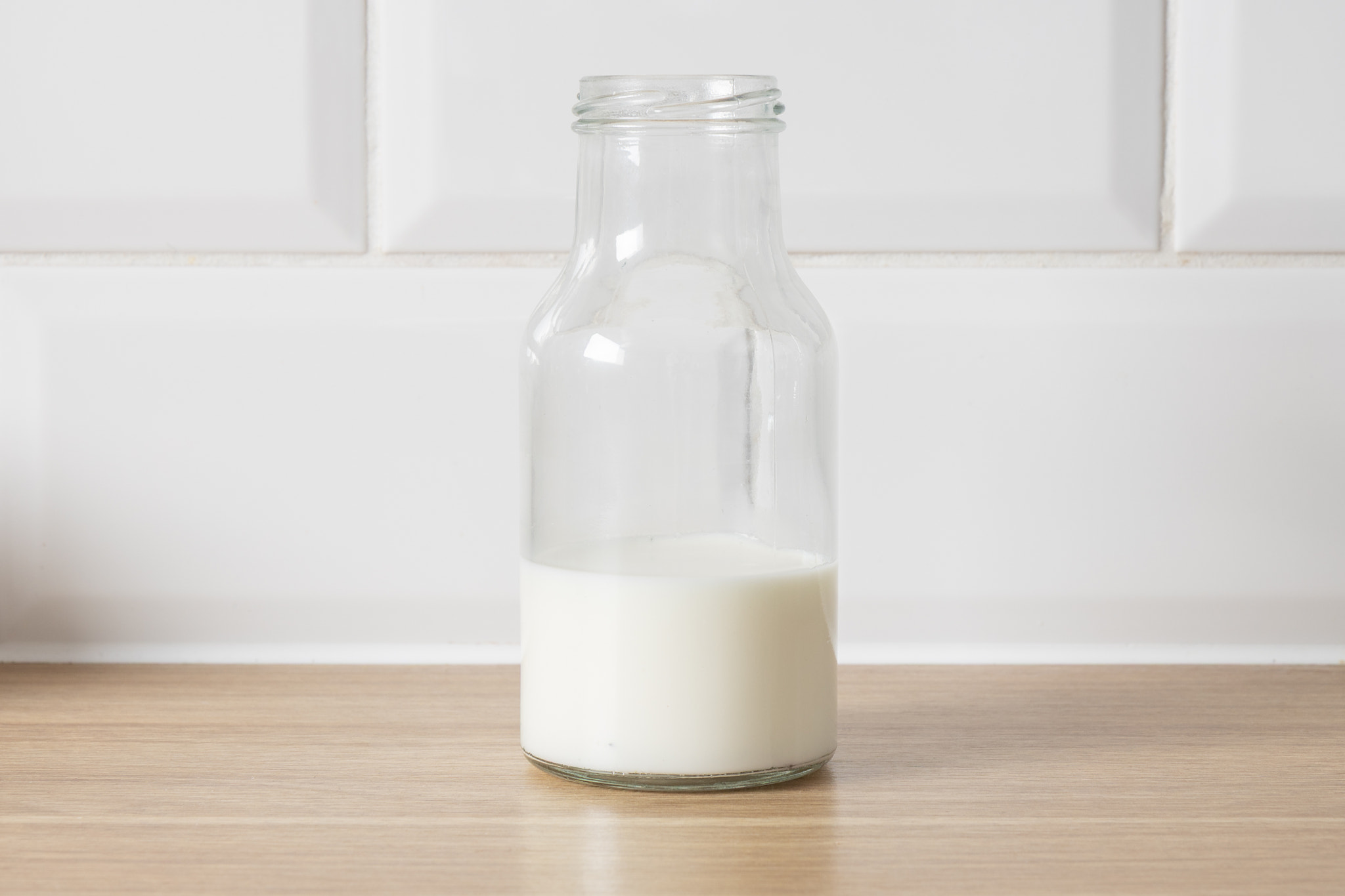 A clear glass bottle filled with milk sits on a wooden countertop against a white tiled wall.