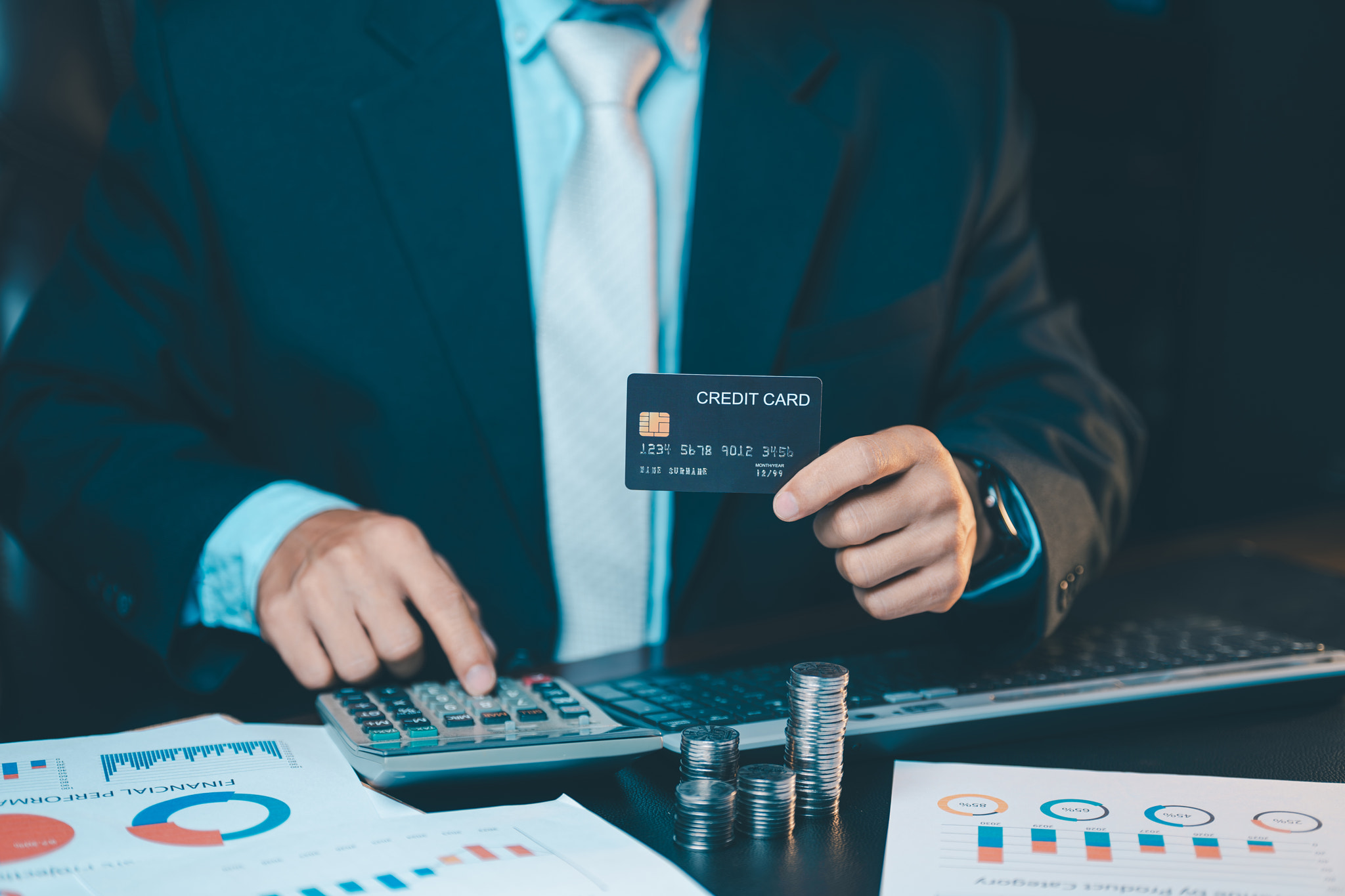 Close up of businessman hand holding a black credit card and using calculator with coins stack on ta