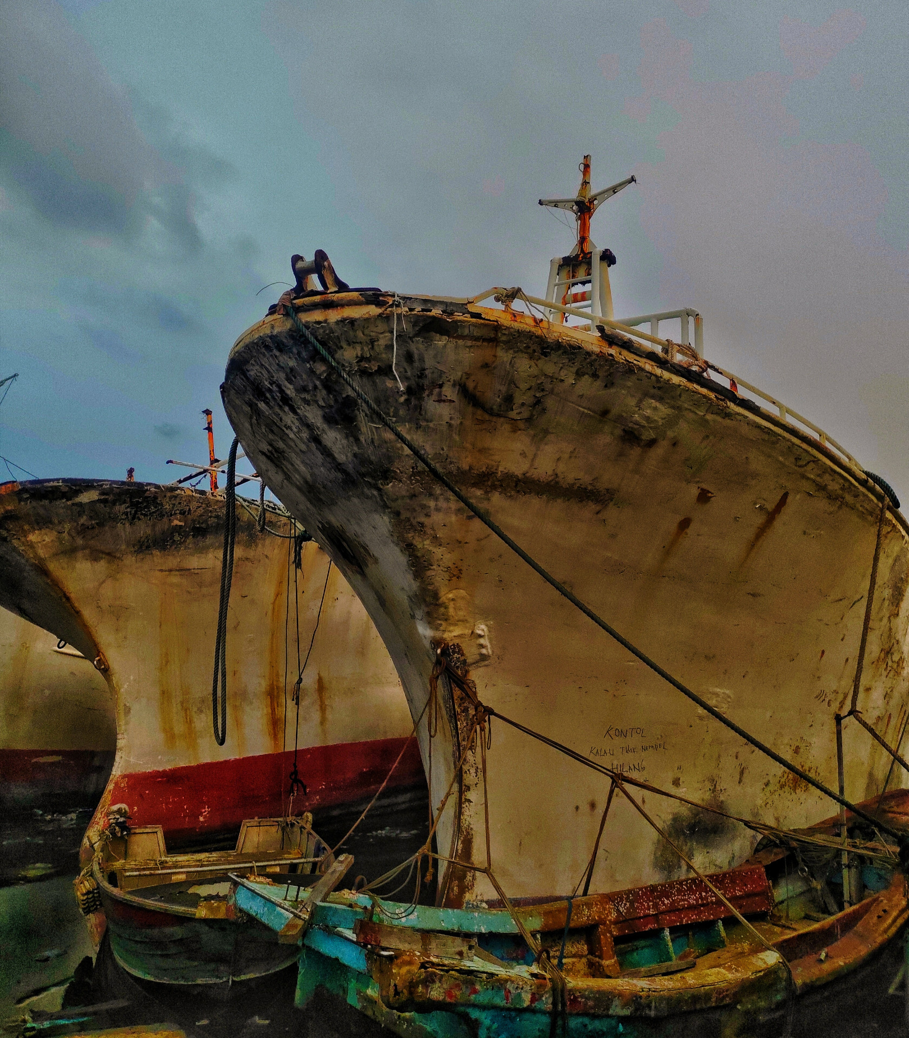 THE MOURNERS.: He didn＇t die alone. They lined up behind him. Hull after hull. Rust after rust. A funeral without a priest. The small boats? Offerings from people who couldn＇t afford flowers. Look closer. Someone carved his rage on the hull: ＂KONTOL KALAU GAK NEMPEL HILANG.＂ Dark Indonesian comedy. A final prayer from a world that left them here to rot. This isn＇t a shipyard. This is a family cemetery in North Jakarta＇s old harbor. Where steel learns to grieve in silence. tool: silence