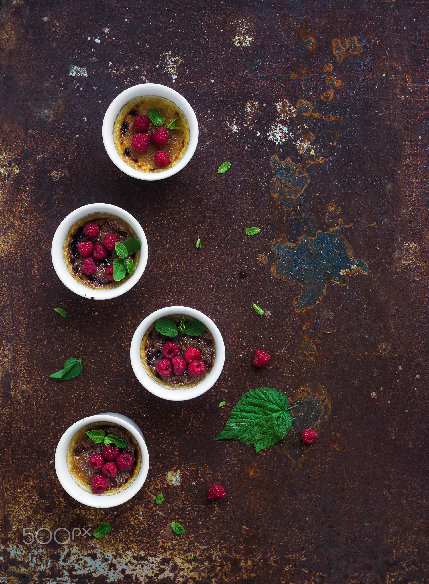 Creme brulees with raspberries and mint in white bowls over grunge metal backdrop. Top view