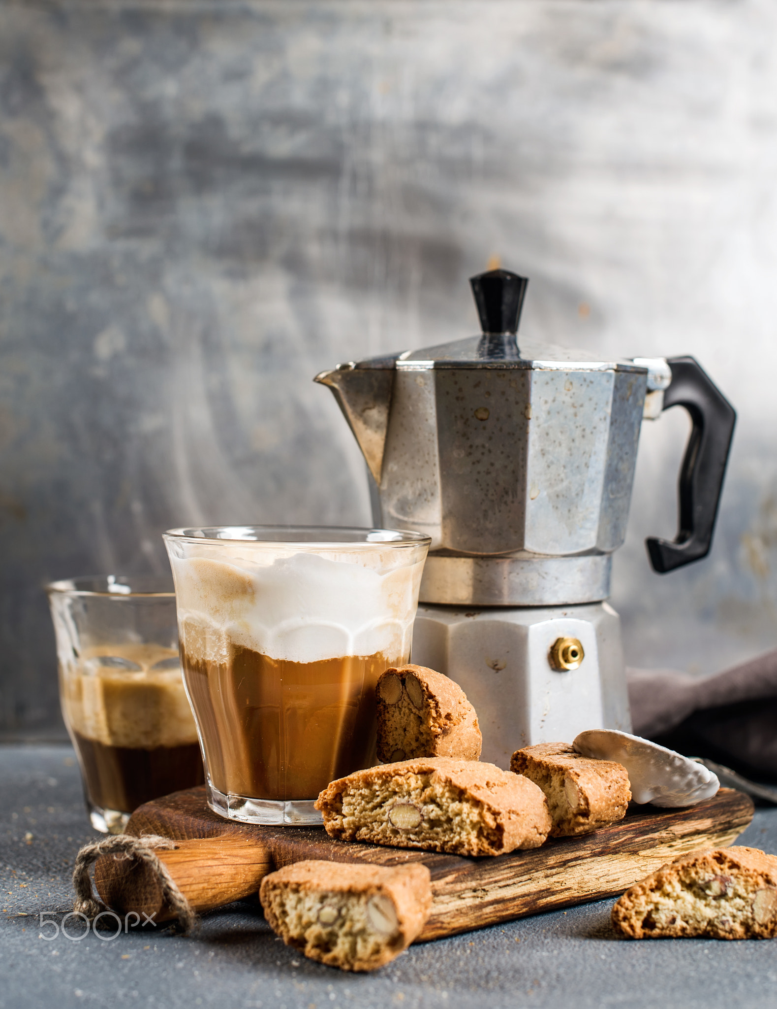 Glass of latte coffee on rustic wooden board, cantucci biscuits and steel Italian Moka pot, grey..