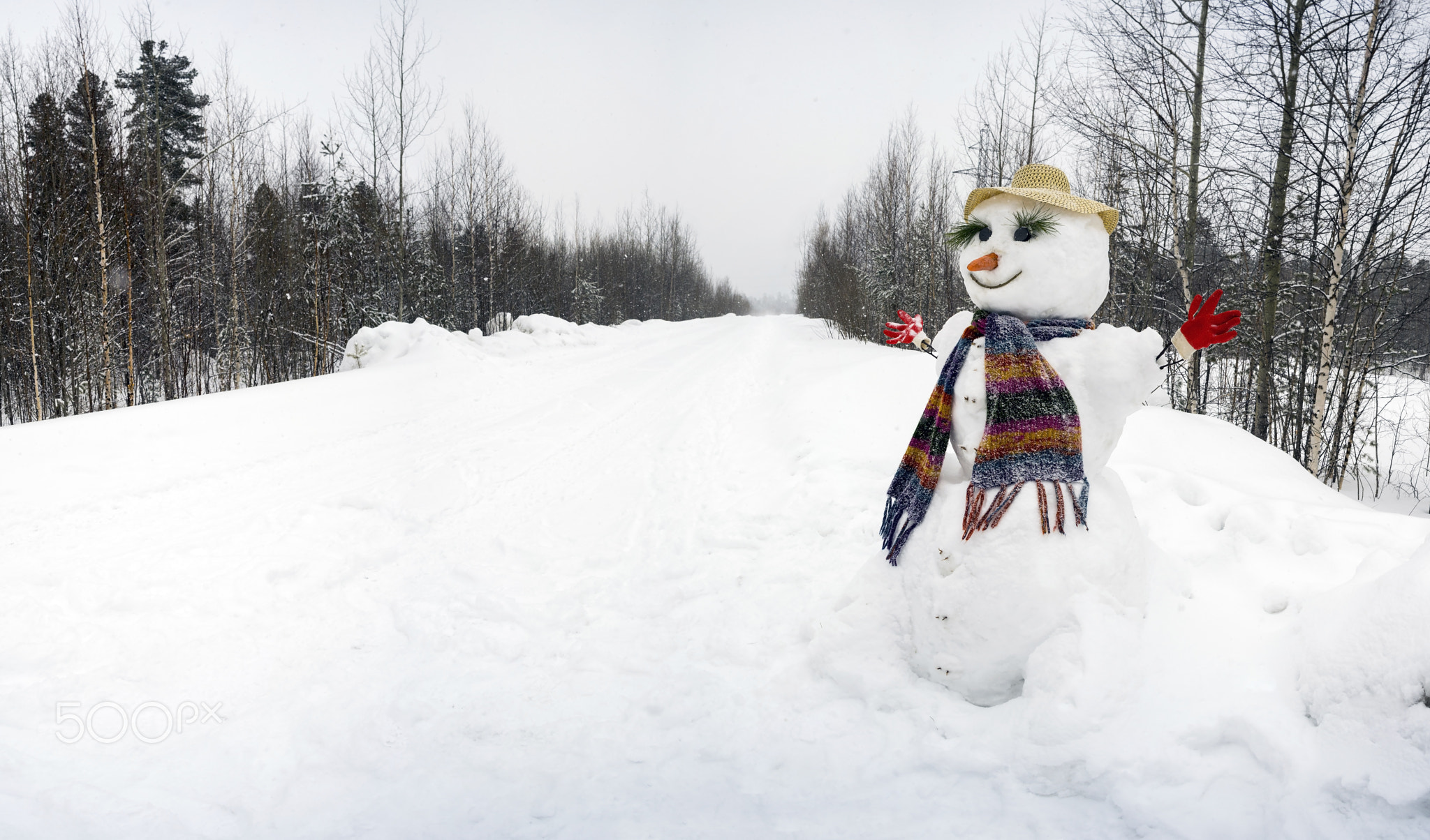 The snowman stands alone on  edge of the road.