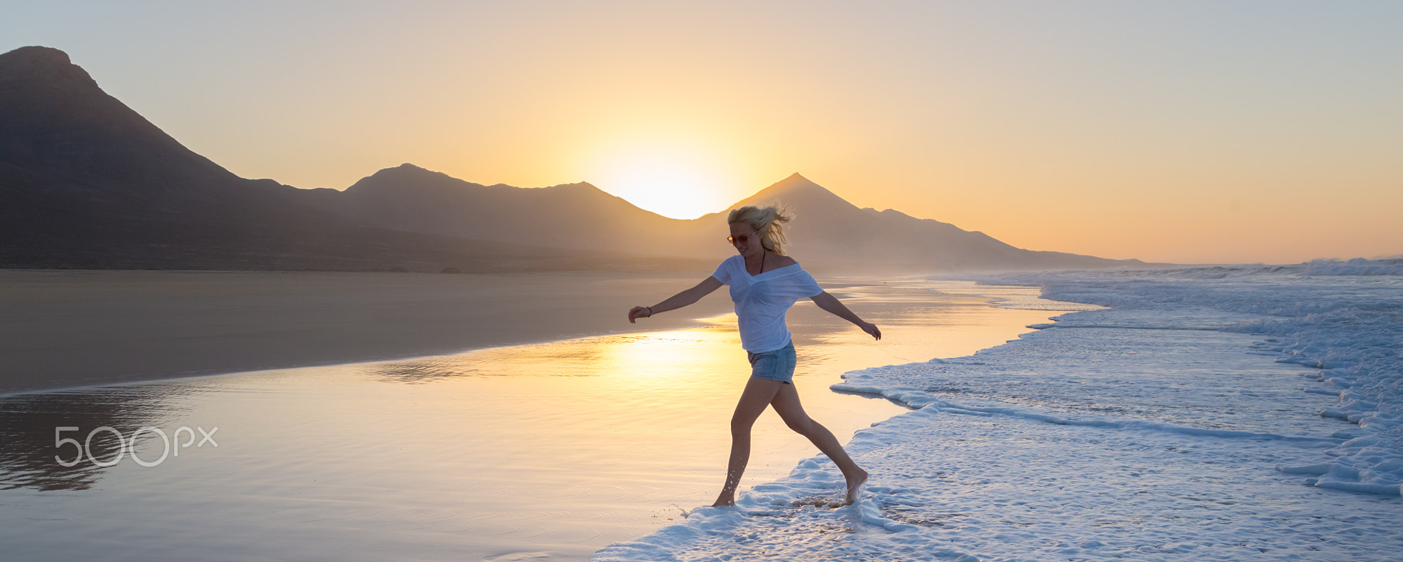 Lady enjoying running from waves on sandy beach in sunset.