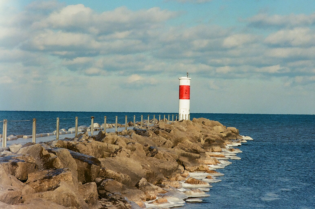 On a partly cloudy day, a rocky pier juts out into the deep blue water. At the end of the pier sits a stout red-and-white lighthouse. The path, as well as the rocks, are covered in ice.
