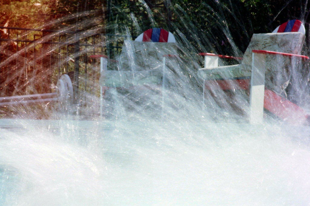 Two red, white, and blue Adirondack chairs. In front, splashes of poolwater are suspended like cellophane.