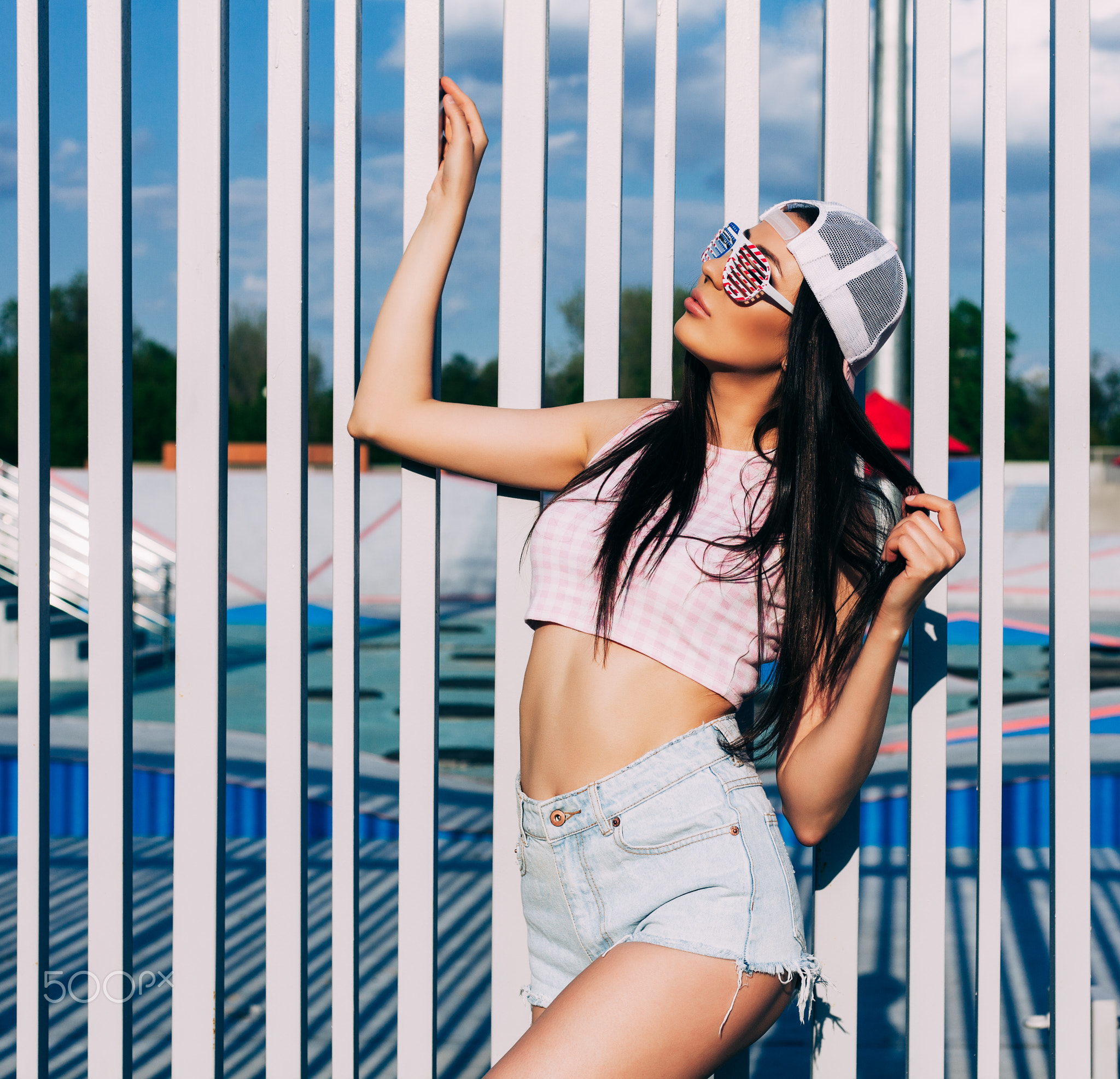 Amazing brunette woman posing in pink top, jeans shorts, baseball cap and trendy sunglasses in...