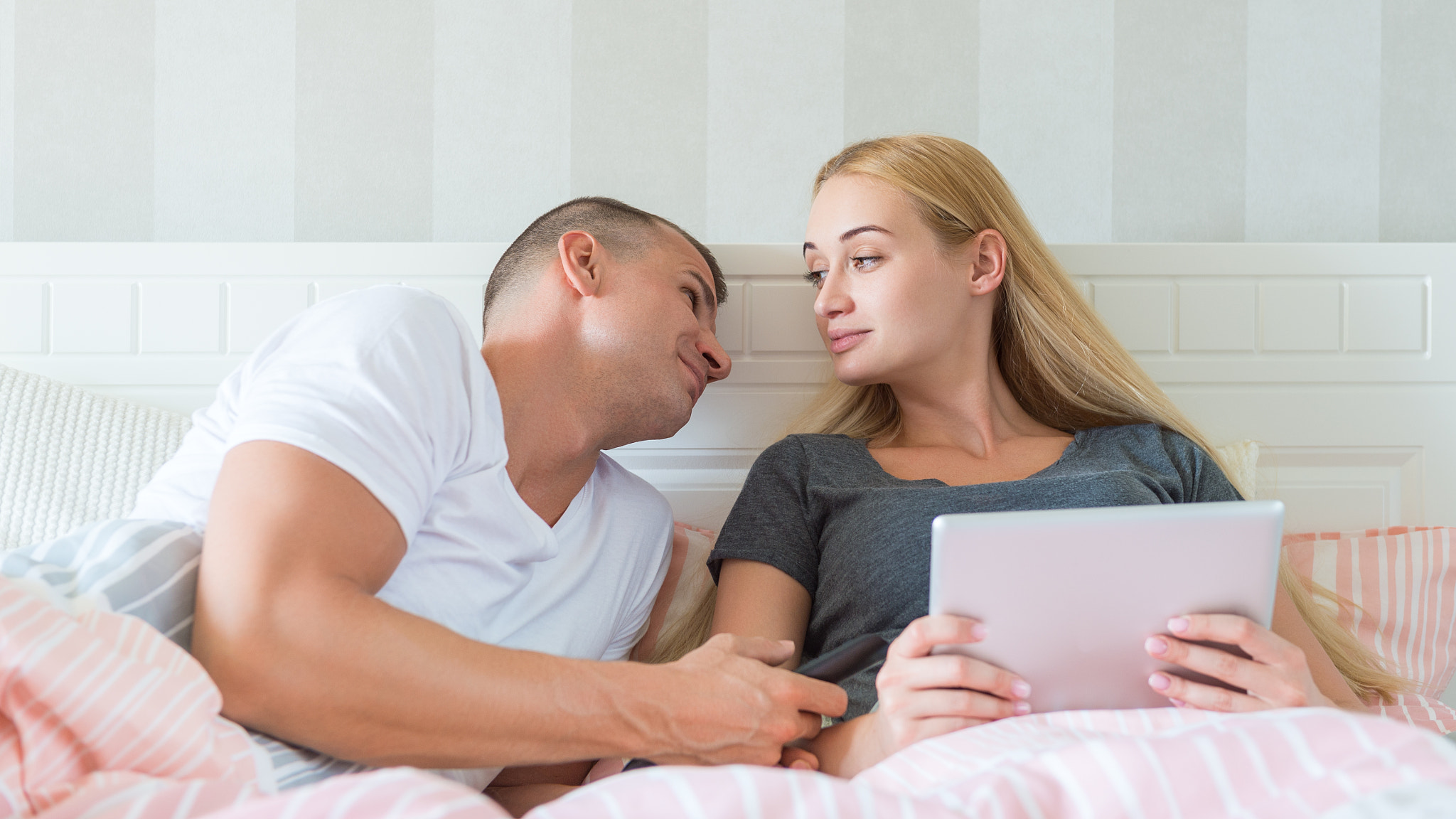 Young attractive caucasian couple in bed. Woman holding digital tablet, man lovingly looking at...