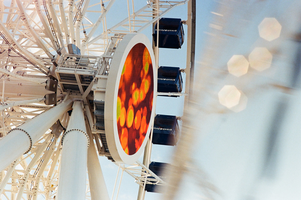 Close-up of a spinning white Ferris wheel. Four deep blue cabins are adorned with the words “Navy Pier”. A large, circular LED screen is attached to the face of the Ferris wheel, displaying orange bokeh. There is photographic white bokeh and a motion blur in front of the scene.