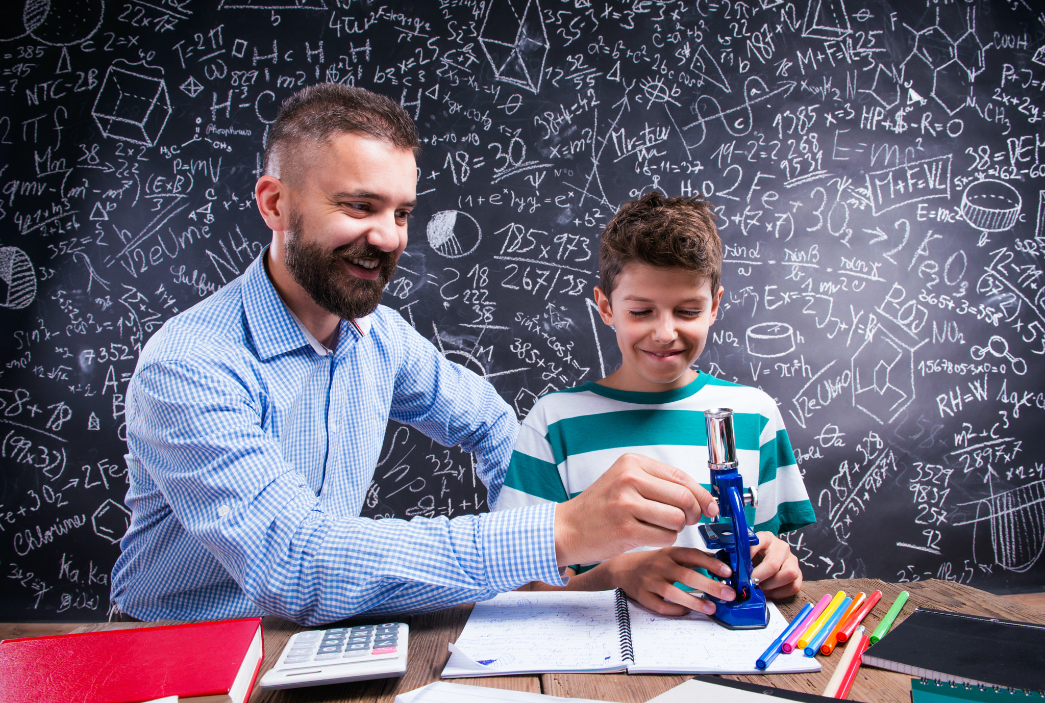 Hipster teacher with his student with microscope, big blackboard