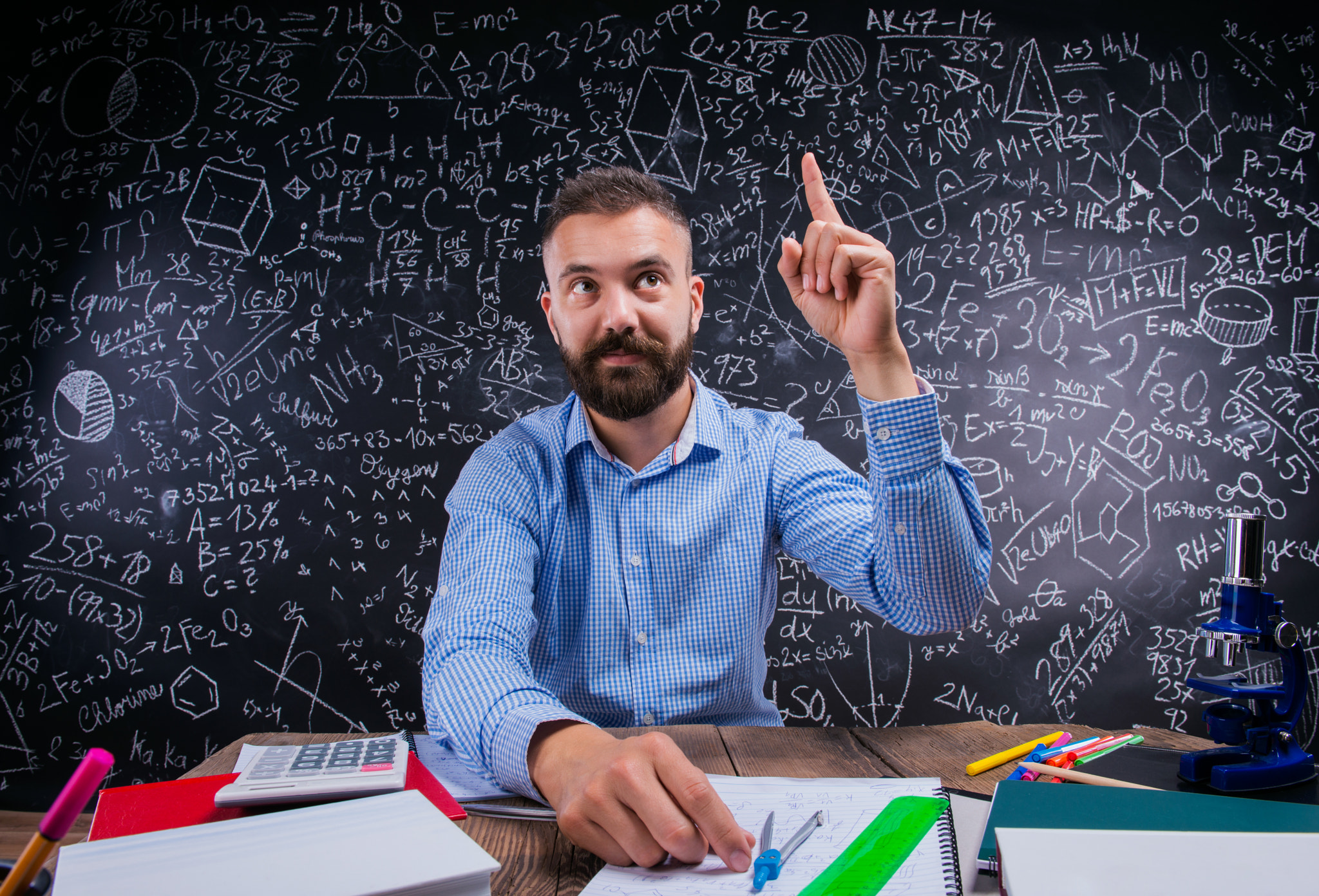 Teacher at desk, school supplies, raised finger, big blackboard